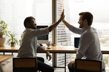 Happy two diverse male employees giving high five, making common decision negotiating online business project, developing growth corporate strategy, sitting together at shared table in modern office.