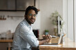 © fizkes - Portrait of happy handsome young african american hipster man in eyeglasses sitting at table with computer. Smiling brazilian businessman professional freelance looking at camera, posing in office.