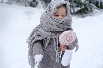 cute baby, girl 2 years old in warm winter retro clothes with caramel candy in winter forest, baby is wrapped in knitted shawl, beautiful landscape, snow-covered fluffy fir trees