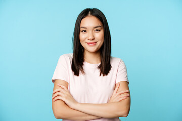 smiling asian girl in t-shirt, holding arms crossed, ready for action pose, standing over blue backg