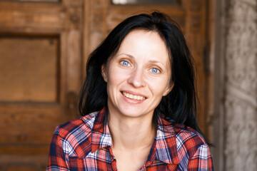 Wall Mural - Close-up face of pretty woman with blue eyes, natural dark hair, no makeup, standing in a plaid shirt smiling, looking at the camera. Against the background of a building with wooden doors