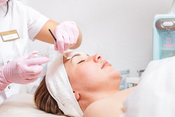 Wall Mural - Beauty salon. The cosmetologist in medical gloves holds a bowl with a mask, applying cosmetics with a brush on the client's face. Copy space. Close up.Concept of professional cosmetology and treatment