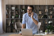 © fizkes - Smiling young successful businessman worker manager in eyewear holding distant phone call conversation with client or partner, standing at table with computer in modern home office workplace.