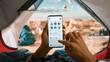© Gorodenkoff - POV of a Tourist Vertically Holding a Smartphone and Checking Social Network Messages, Swiping a Finger Over Display. Traveller Resting in a Tent on Top of a Rocky Mountain and Flying Hot Air Balloons