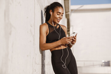 Wall Mural - Black sportswoman listening music while leaning on concrete wall