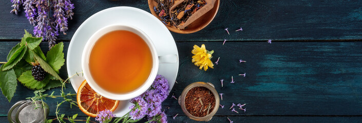 Wall Mural - Tea panorama with a place for text. Herbs, flowers and fruit around a cup of tea, an overhead flat lay shot on a dark rustic wooden background