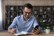 © fizkes - Happy young handsome business man in eyeglasses looking at cellphone screen, web surfing useful information writing notes in paper planner, planning workday or checking working schedule at home office
