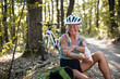 © Halfpoint - Happy senior woman biker sitting and applying sunscreen on her arm outdoors in forest.