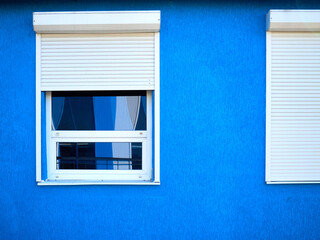 two windows of a part of the building, white shutters, blinds are closed at one window, there is a reflection of the street in the other window, the walls of the building are painted blue