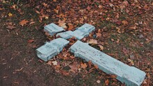 Devastated Shattered Stone Concrete Crucifix In Act Of Religious Hatred And Persecution Left On Ground Amoung Withered Autumn Foliage Leaves