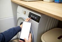 Woman Operating Washing Machine Through Smart Phone In Utility Room At Home