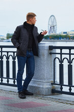 A Young Man Smokes A Pipe On The City Embankment. Ferris Wheel In The Background Behind The River In Blur.