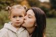 © Ananass - Young happy and smiling mom with her little daughter in arms hugging and kissing spending a weekend on a walk in autumn park. selective focus, noise effect, Autumnal mood