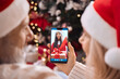 © insta_photos - Happy senior grandparents couple in santa hats greeting grandchild opening Christmas gift on family cell phone video call, online virtual party meeting videocall on smartphone at home. Over shoulder