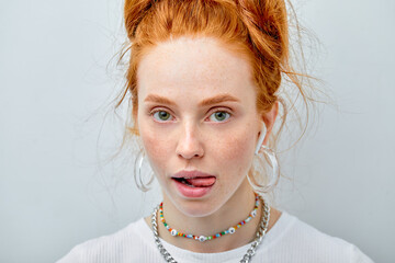 Wall Mural - Portrait of cheerful playful young woman with red hair in bun showing tongue isolated over white studio background. Cool freckled caucasian lady in headphones look at camera charmingly, sexy