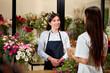 Side View On Young Friendly Woman Florist Having Talk With Customer Client In Shop. Smiling Female In Apron At Work, Consult And Serve Client. Different Various Bouquets in The Background