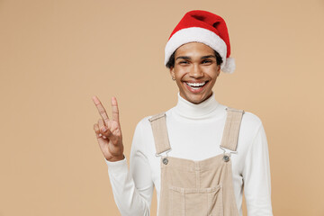 Wall Mural - Young cheerful smiling happy african man 20s wear Santa Claus red Christmas hat showing victory sign isolated on plain pastel beige background studio portrait. Happy New Year 2022 celebration concept.