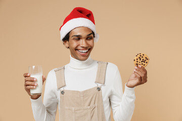 Wall Mural - Young cheerful fun african man 20s in Santa Claus red Christmas hat eat cookies biscuits drink milk isolated on plain pastel beige background studio portrait. Happy New Year 2022 celebration concept.