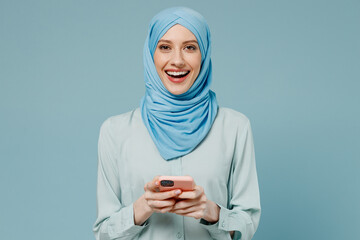 Young amazed happy arabian asian muslim woman in abaya hijab hold in hand use mobile cell phone isolated on plain blue background studio portrait. People uae middle eastern islam religious concept