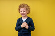 smiling Cute caucasian kid boy holding hands on chest near heart, casually dressed, having curly hair. Human emotions, real feelings and facial expression concept. isolated on yellow background