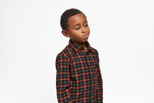 Horizontal Studio Picture Of Upset Cute Dark-skinned Boy In Plaid Red And Black Shirt Looking Aside With Tighten Lips And Disappointed Face Expression. Children. Negative Human Emotions