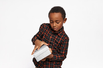 Wall Mural - Studio portrait of handsome preschool boy with dark skin opening silver box with gift or present, holding it carefully, wearing red flannel shirt isolated on white wall with copy space for your text