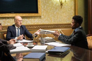 Aufkleber - Young African male lawyer in formalwear passing folder with papers to mature colleague over table with other documents