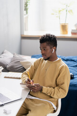 Poster - Serious young African student making notes in notepad while sitting by desks in front of computer