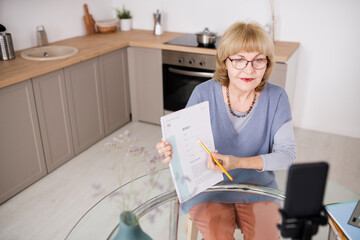 Aufkleber - Confident mature teacher pointing at paper while sitting by kitchen table in front of smartphone camera during livestream