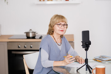 Poster - Confident mature female coach looking in smartphone camera during online training while sitting by kitchen table