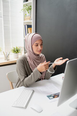 Aufkleber - Young confident teacher in hijab speaking to online audience in front of computer monitor while sitting by desk in home environment