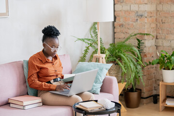 Poster - Young African woman with laptop on knees sitting on sofa in living-room during online lesson and looking through lecture notes