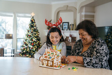Girl And Grandma Decorating Gingerbread House In Front Of Christmas Tree