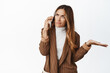 Confused businesswoman talking on cellphone, shrugging while listening caller, standing in brown suit over white background