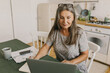 © shurkin_son - Good-looking elderly grey-haired woman in cute dress sitting in kitchen in front of laptop studying online, taking notes after her teacher, watching on screen with enthusiasm and happy face