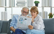 © Bangkok Click Studio - Caucasian old senior elderly grandparent grey bearded hair husband and pretty wife sitting cuddling together on cozy sofa in living room at home in morning reading funny news on daily newspaper