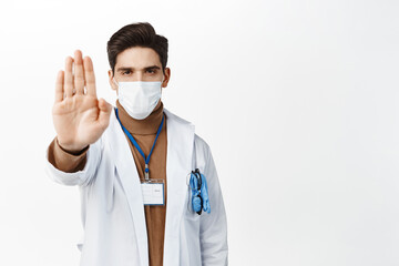 Wall Mural - Covid-19, quarantine and healthcare concept. Serious doctor in robe and face mask, showing stop sign, extend one palm in disapproval, reject smth, standing over white background