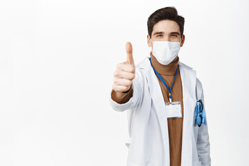 Wall Mural - Coronavirus and vaccination concept. Young doctor in medical face mask and robe, showing thumb up, approve, like smth good, standing over white background