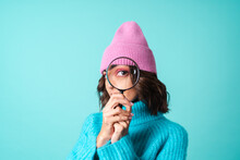 Cozy Portrait Of A Young Woman In A Knitted Blue Sweater And A Pink Hat With Bright Makeup Holding A Magnifying Glass, Fooling Around, Having Fun