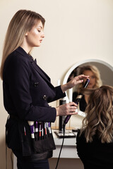 Wall Mural - hair master makes curls with curling iron for young woman with blond hair. Reflection in dressing room mirror.