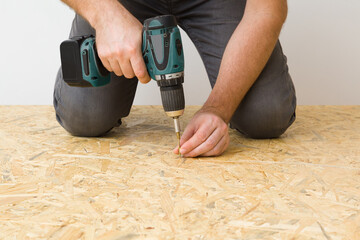 Young adult man hands using battery screwdriver and screwing screw in osb board for floor. Closeup. Front view. Repair work of home. Renovation process.