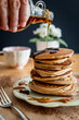 Hand pouring maple syrup on a healthy buckwheat pancake stack topped with chocolate and halvah on a decorative plate. Easy to make gluten free morning breakfast, brunch. Delicious golden pancake heap.