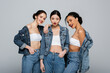 © LIGHTFIELD STUDIOS - Smiling interracial women in denim jackets looking at camera isolated on grey