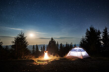 White Tent Locating On Mountain Hill Near Burning Campfire At Forest. Moonlight In The Magic Starry Sky Overnight Camping In The Mountains. Concept Of Travelling, Hiking And Night Camping.