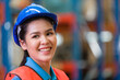 © amorn - Head shot of Asian woman worker wearing safety vest and helmet at work in the industry storage warehouse. Smiling female worker at warehouse. Inspection quality control