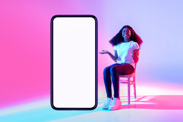 Wall Mural - Lovely black lady sitting on chair near modern smartphone with empty screen and offering space for mockup in neon light