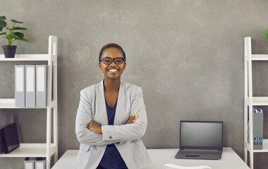 Sticker - Portrait of happy smiling black businesswoman at work. Beautiful young African American business woman in jacket and glasses standing arms folded leaning on table in office with grey copy space wall