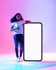 Poster - Happy black woman standing near big smartphone with blank white screen, gesturing thumb up in neon light, mockup