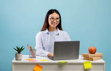 Wall Mural - Happy asian lady using laptop while learning online at desk over blue background, smiling to camera