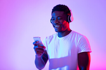 Poster - Cheerful black guy in wireless headphones using smartphone in neon light